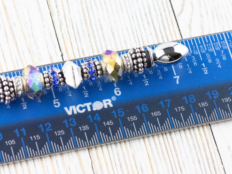 K&ouml;nnte beinhalten: Nahaufnahme eines silbernen Charm-Armbands mit verschiedenen Perlen, darunter eine klare, schillernde Perle, eine blaue Kristallperle, eine silberne Perle mit strukturierter Oberfl&auml;che, eine klare Kristallperle mit goldfarbenem Akzent, eine silberne Perle mit strukturierter Oberfl&auml;che und eine silberne Perle mit strukturierter Oberfl&auml;che. Das Armband wird auf einem blauen Lineal mit schwarzen Markierungen gemessen.