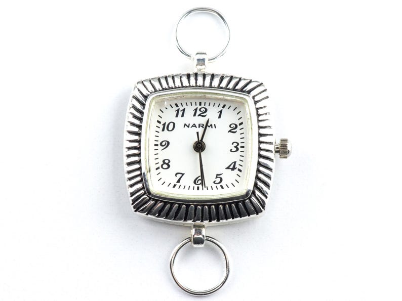 May include: A silver pocket watch with a square face and a white dial. The watch has a textured bezel and a small crown. The dial features black numerals and the brand name "NARMI". Two silver rings are attached to the top and bottom of the watch.