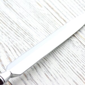 May include: A shiny, silver letter opener with a pointed blade. The handle has a decorative, geometric design. The opener is set against a light-colored, textured wooden surface.