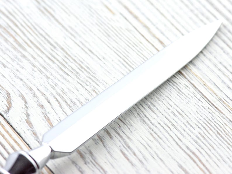 May include: A shiny, silver letter opener with a pointed blade. The handle has a decorative, geometric design. The opener is set against a light-colored, textured wooden surface.