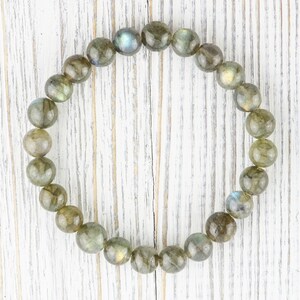 Puede incluir: Una pulsera redonda hecha de cuentas de labradorita pulidas. Las cuentas son una mezcla de gris, verde y marr&oacute;n con destellos iridiscentes de azul y dorado. La pulsera se exhibe sobre una superficie de madera blanca.