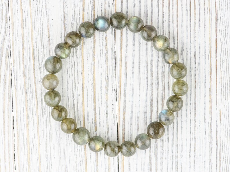 Puede incluir: Una pulsera redonda hecha de cuentas de labradorita pulidas. Las cuentas son una mezcla de gris, verde y marr&oacute;n con destellos iridiscentes de azul y dorado. La pulsera se exhibe sobre una superficie de madera blanca.