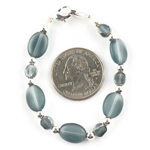 Pu&ograve; includere: Un braccialetto di perline con perline ovali grigio-blu, perline sfaccettate pi&ugrave; piccole e dettagli in metallo color argento. Il braccialetto &egrave; mostrato accanto a una moneta per il confronto delle dimensioni. Il braccialetto ha una chiusura argentata.