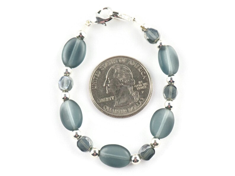 Pu&ograve; includere: Un braccialetto di perline con perline ovali grigio-blu, perline sfaccettate pi&ugrave; piccole e dettagli in metallo color argento. Il braccialetto &egrave; mostrato accanto a una moneta per il confronto delle dimensioni. Il braccialetto ha una chiusura argentata.