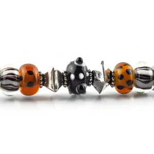 K&ouml;nnte beinhalten: Ein silbernes Armband mit verschiedenen bunten Glasperlen, darunter schwarz-wei&szlig;e Tupfen, orange mit schwarzen Punkten und wei&szlig; mit schwarzen Streifen.
