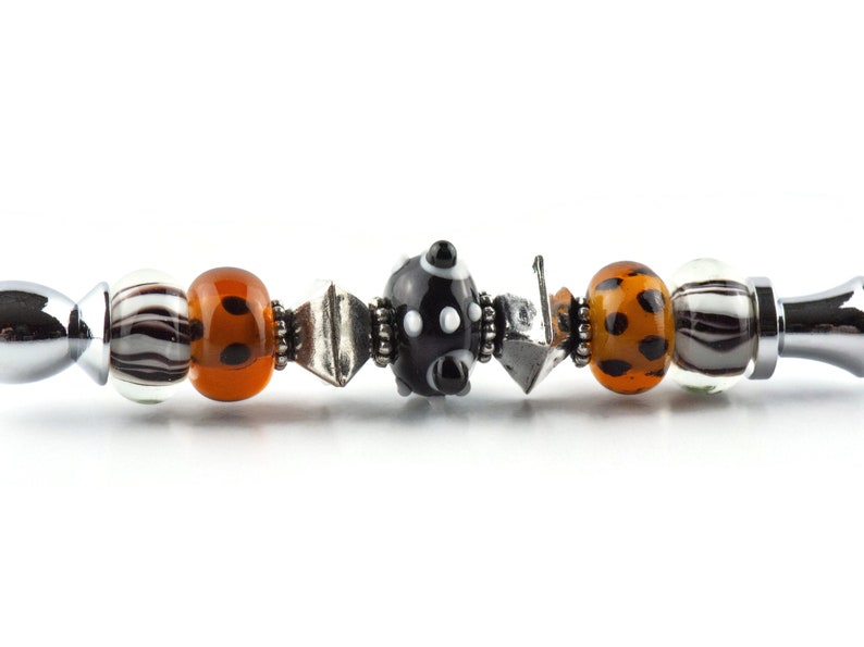K&ouml;nnte beinhalten: Ein silbernes Armband mit verschiedenen bunten Glasperlen, darunter schwarz-wei&szlig;e Tupfen, orange mit schwarzen Punkten und wei&szlig; mit schwarzen Streifen.