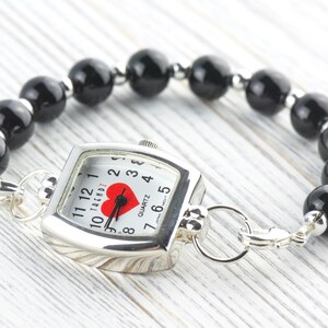 Puede incluir: Un reloj plateado con esfera cuadrada y pulsera de cuentas negras. La esfera del reloj tiene un coraz&oacute;n rojo y manecillas negras. La pulsera est&aacute; hecha de cuentas negras redondas.