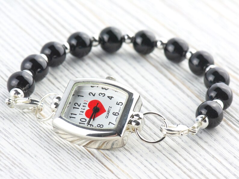 Puede incluir: Un reloj plateado con esfera cuadrada y pulsera de cuentas negras. La esfera del reloj tiene un coraz&oacute;n rojo y manecillas negras. La pulsera est&aacute; hecha de cuentas negras redondas.