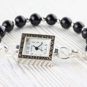 Peut inclure: Une montre Geneva &agrave; quartz avec un cadran carr&eacute; argent&eacute; et une bordure dor&eacute;e d&eacute;corative. La montre est attach&eacute;e &agrave; un bracelet compos&eacute; de grosses perles noires rondes et de fermoirs argent&eacute;s.