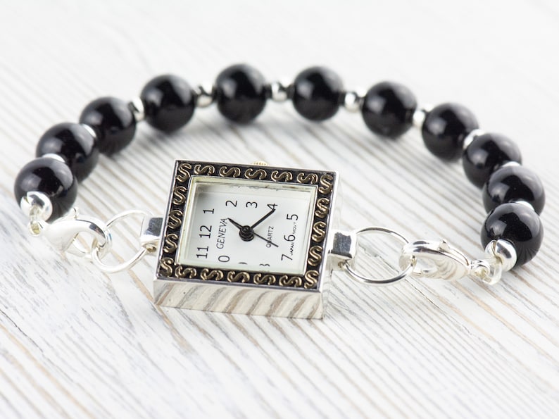 Peut inclure: Une montre Geneva &agrave; quartz avec un cadran carr&eacute; argent&eacute; et une bordure dor&eacute;e d&eacute;corative. La montre est attach&eacute;e &agrave; un bracelet compos&eacute; de grosses perles noires rondes et de fermoirs argent&eacute;s.