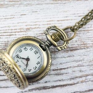 K&ouml;nnte beinhalten: Antike bronzefarbene Taschenuhr mit wei&szlig;em Zifferblatt und schwarzen Ziffern. Die Uhr hat einen offenen Deckel, der die Zeit anzeigt. Die Uhr ist an einer passenden Bronzekette befestigt. Der Hintergrund ist eine wei&szlig;e Holzoberfl&auml;che.