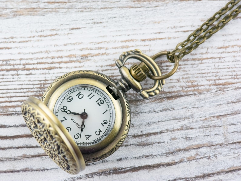 K&ouml;nnte beinhalten: Antike bronzefarbene Taschenuhr mit wei&szlig;em Zifferblatt und schwarzen Ziffern. Die Uhr hat einen offenen Deckel, der die Zeit anzeigt. Die Uhr ist an einer passenden Bronzekette befestigt. Der Hintergrund ist eine wei&szlig;e Holzoberfl&auml;che.