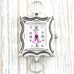 K&ouml;nnte beinhalten: Eine silberfarbene Geneva-Quarzuhr mit wei&szlig;em Zifferblatt, schwarzen Ziffern und einem rosa Schleifendesign. Die Uhr hat einen quadratischen, dekorativen Rahmen und einen Ring oben und unten. Die Uhr ist ein Symbol f&uuml;r Brustkrebsbewusstsein.