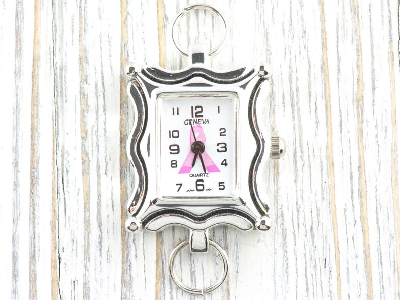 K&ouml;nnte beinhalten: Eine silberfarbene Geneva-Quarzuhr mit wei&szlig;em Zifferblatt, schwarzen Ziffern und einem rosa Schleifendesign. Die Uhr hat einen quadratischen, dekorativen Rahmen und einen Ring oben und unten. Die Uhr ist ein Symbol f&uuml;r Brustkrebsbewusstsein.