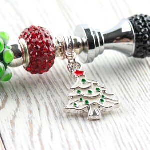 May include: A festive charm bracelet with a Christmas theme. It features a silver charm with a white Christmas tree design, red and green beads, and a black crystal bead. The bracelet is set against a white wooden background.