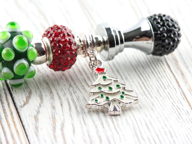 May include: A festive charm bracelet with a Christmas theme. It features a silver charm with a white Christmas tree design, red and green beads, and a black crystal bead. The bracelet is set against a white wooden background.