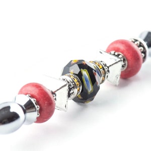 May include: A decorative pen featuring a silver metal body with red, black, and multi-colored beads. The pen has a faceted black bead with colorful accents, red round beads, and silver square and round accents. The pen is on a white background.