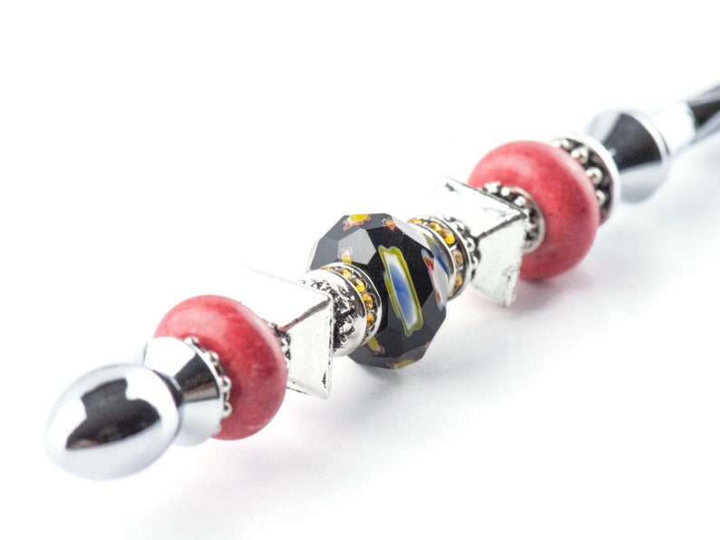 May include: A decorative pen featuring a silver metal body with red, black, and multi-colored beads. The pen has a faceted black bead with colorful accents, red round beads, and silver square and round accents. The pen is on a white background.