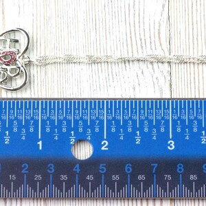 Pode incluir: Colar prateado com um pendente em forma de cora&ccedil;&atilde;o. O pendente apresenta a palavra "HOPE" com uma fita rosa e um pequeno desenho. A corrente do colar tem aproximadamente 25 cm de comprimento. Uma r&eacute;gua azul est&aacute; em primeiro plano.