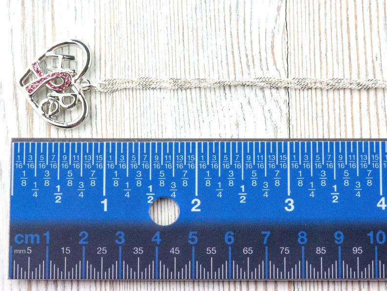 Pode incluir: Colar prateado com um pendente em forma de cora&ccedil;&atilde;o. O pendente apresenta a palavra "HOPE" com uma fita rosa e um pequeno desenho. A corrente do colar tem aproximadamente 25 cm de comprimento. Uma r&eacute;gua azul est&aacute; em primeiro plano.
