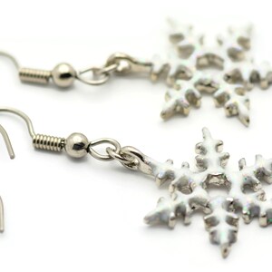 May include: A pair of silver snowflake earrings with white enamel. The earrings have a hook closure.