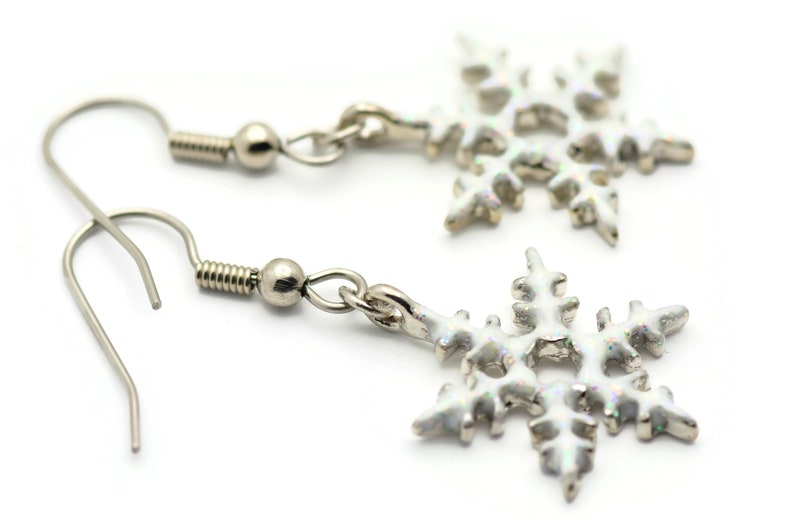 May include: A pair of silver snowflake earrings with white enamel. The earrings have a hook closure.