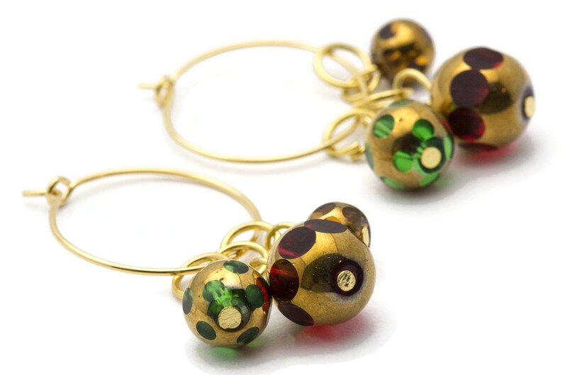 May include: Gold hoop earrings with dangling charms. The charms are round and have a gold finish with green and red patterned glass.