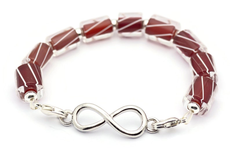 K&ouml;nnte beinhalten: Ein Armband aus roten und wei&szlig;en, gestreiften Glasperlen mit einem Unendlichkeits-Anh&auml;nger. Das Armband ist aus Silber und hat einen Verschluss.