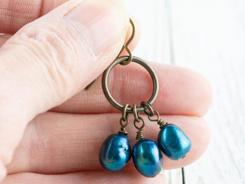 K&ouml;nnte beinhalten: Ohrringe mit einem bronzefarbenen Metallring und Haken. Drei tropfenf&ouml;rmige, irisierende blaue Perlen h&auml;ngen an dem Ring. Die Ohrringe werden in einer Hand gehalten.
