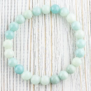 May include: A light blue and green beaded bracelet made of round, smooth stones. The bracelet is laid out in a circle on a white wooden surface.