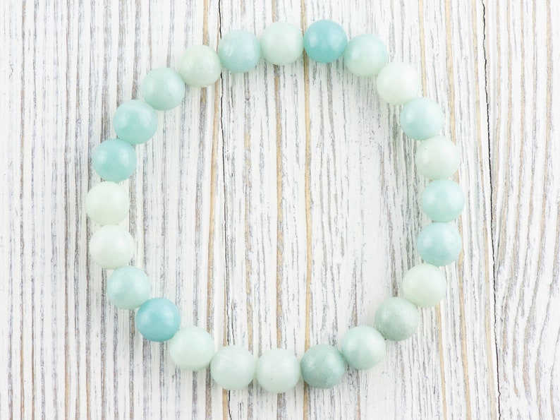 May include: A light blue and green beaded bracelet made of round, smooth stones. The bracelet is laid out in a circle on a white wooden surface.