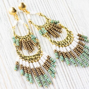 May include: A pair of long, gold hoop earrings with white and green beads hanging from the bottom. The earrings have a hammered metal finish.