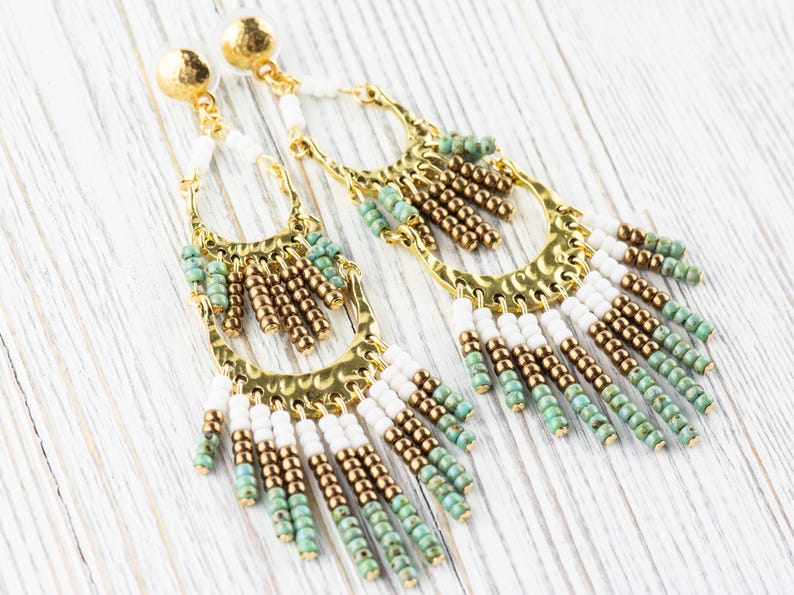 May include: A pair of long, gold hoop earrings with white and green beads hanging from the bottom. The earrings have a hammered metal finish.