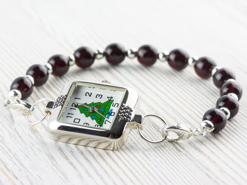 Puede incluir: Reloj plateado con esfera cuadrada con dise&ntilde;o de &aacute;rbol de Navidad. La correa est&aacute; hecha de cuentas rojas oscuras y detalles plateados. Un accesorio festivo para las fiestas.