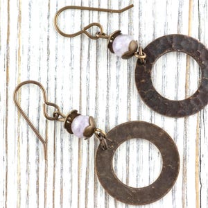 Puede incluir: Un par de pendientes hechos a mano con aros de metal color bronce martillado, una peque&ntilde;a cuenta de color p&uacute;rpura claro y ganchos para las orejas de color bronce. Los pendientes se exhiben sobre una superficie de madera blanca.