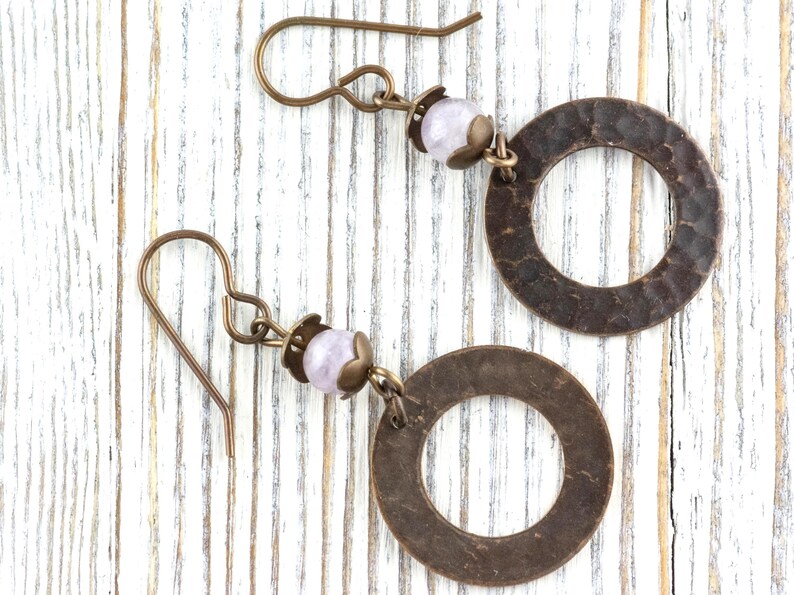 Puede incluir: Un par de pendientes hechos a mano con aros de metal color bronce martillado, una peque&ntilde;a cuenta de color p&uacute;rpura claro y ganchos para las orejas de color bronce. Los pendientes se exhiben sobre una superficie de madera blanca.