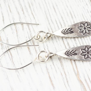 Puede incluir: Un par de pendientes de plata con un dije en forma de l&aacute;grima que presenta un dise&ntilde;o de flor y tres marcas en forma de flecha.