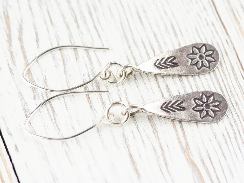 Puede incluir: Un par de pendientes de plata con un dije en forma de l&aacute;grima que presenta un dise&ntilde;o de flor y tres marcas en forma de flecha.