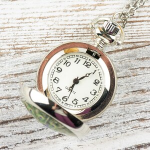 May include: A silver pocket watch with a white face and black hands. The watch has a peacock design on the back. The watch is open and the face is visible. The watch is on a white wooden surface.