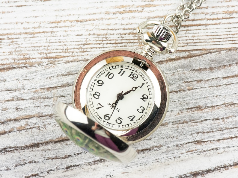 May include: A silver pocket watch with a white face and black hands. The watch has a peacock design on the back. The watch is open and the face is visible. The watch is on a white wooden surface.
