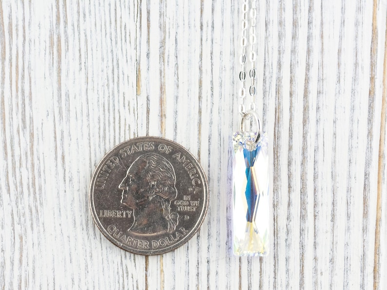 Pu&ograve; includere: Una collana con catena d'argento con un ciondolo di cristallo trasparente. Il ciondolo &egrave; rettangolare e presenta un design sfaccettato. La collana &egrave; mostrata accanto a un quarto di dollaro americano per riferimento alle dimensioni.