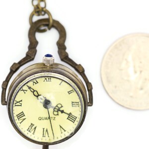 May include: A gold-toned pocket watch with a Roman numeral clock face and a blue stone on the winding stem. The watch is attached to a chain and is about the size of a UK 20 pence coin.