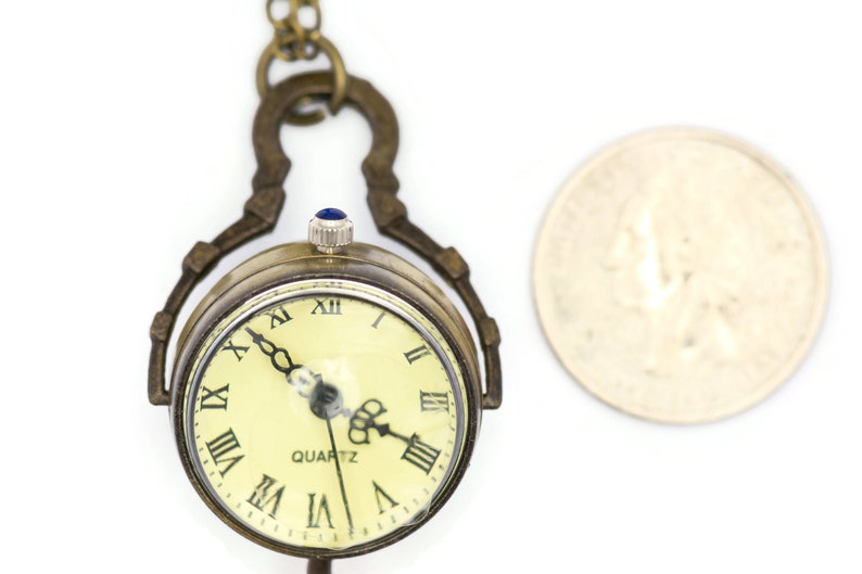 May include: A gold-toned pocket watch with a Roman numeral clock face and a blue stone on the winding stem. The watch is attached to a chain and is about the size of a UK 20 pence coin.