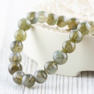 Puede incluir: Un brazalete de cuentas con grandes cuentas redondas de labradorita. Las cuentas muestran una mezcla de tonos grises, verdes y dorados. El brazalete se exhibe sobre una superficie blanca, resaltando los colores naturales de las piedras.