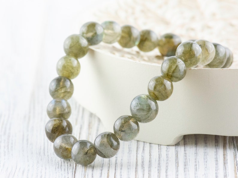 Puede incluir: Un brazalete de cuentas con grandes cuentas redondas de labradorita. Las cuentas muestran una mezcla de tonos grises, verdes y dorados. El brazalete se exhibe sobre una superficie blanca, resaltando los colores naturales de las piedras.
