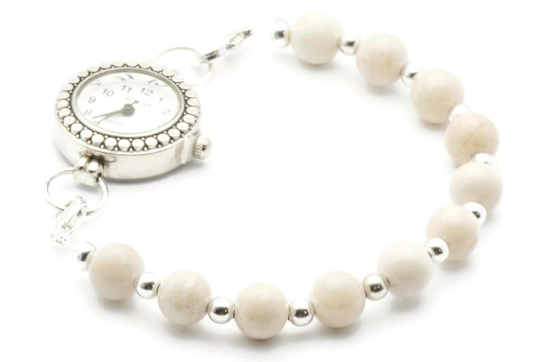 Op de afbeelding: Een zilveren horloge met een witte wijzerplaat en zwarte wijzers is bevestigd aan een armband gemaakt van witte kralen en zilveren tussenstukken.
