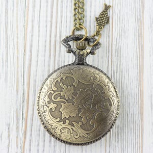 Pode incluir: Um rel&oacute;gio de bolso dourado com uma corrente e um amuleto de peixe. O mostrador do rel&oacute;gio tem um design floral intrincado.