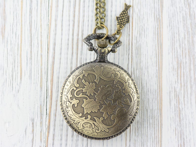 Pode incluir: Um rel&oacute;gio de bolso dourado com uma corrente e um amuleto de peixe. O mostrador do rel&oacute;gio tem um design floral intrincado.