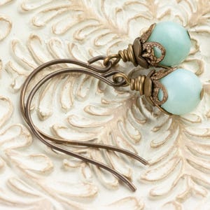 May include: A pair of earrings featuring pale blue, round beads. The beads are accented with ornate, antique bronze-colored metalwork. The ear wires are also bronze-colored and have a simple, curved design. The earrings are displayed on a white, textured surface.