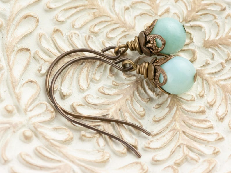 May include: A pair of earrings featuring pale blue, round beads. The beads are accented with ornate, antique bronze-colored metalwork. The ear wires are also bronze-colored and have a simple, curved design. The earrings are displayed on a white, textured surface.